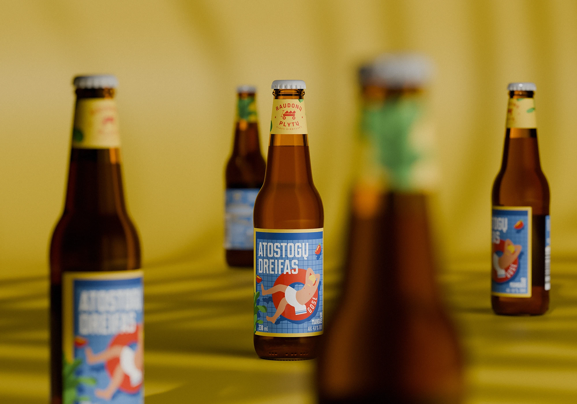 Several brown glass beer bottles with colorful labels stand against a soft yellow background. The central bottle, in sharp focus, features a playful blue label with the name “Atostogų Dreifas” and an illustration of a swimmer in a red swimsuit. The label includes branding for Raudonų Plytų Alaus Dirbtuvės.