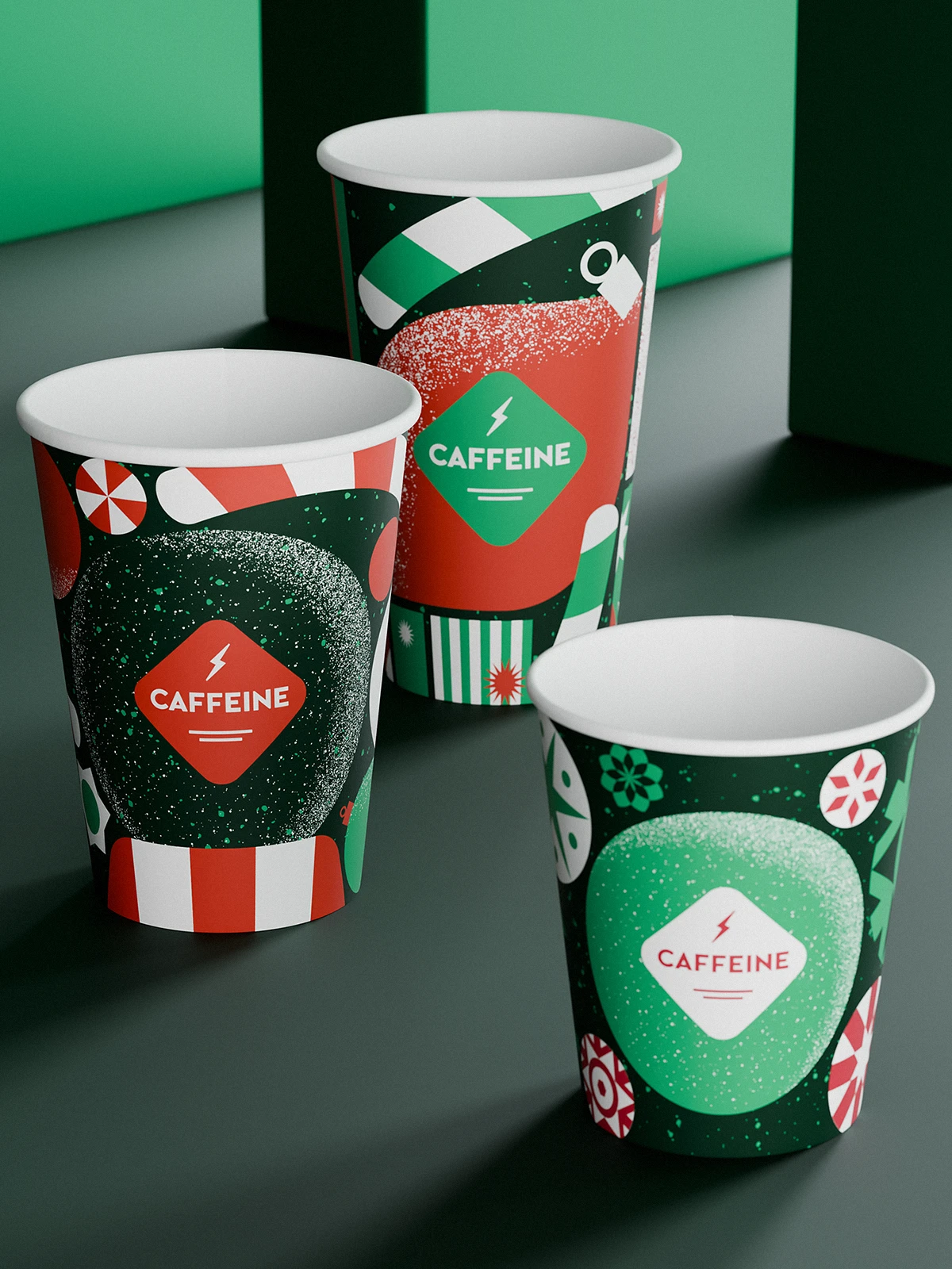 Three CAFFEINE holiday coffee cups with festive 2023 design. The cups feature bold red, green, and white graphics with candy motifs, abstract shapes, and the CAFFEINE logo in diamond frames. Placed on a dark green surface with geometric green background.
