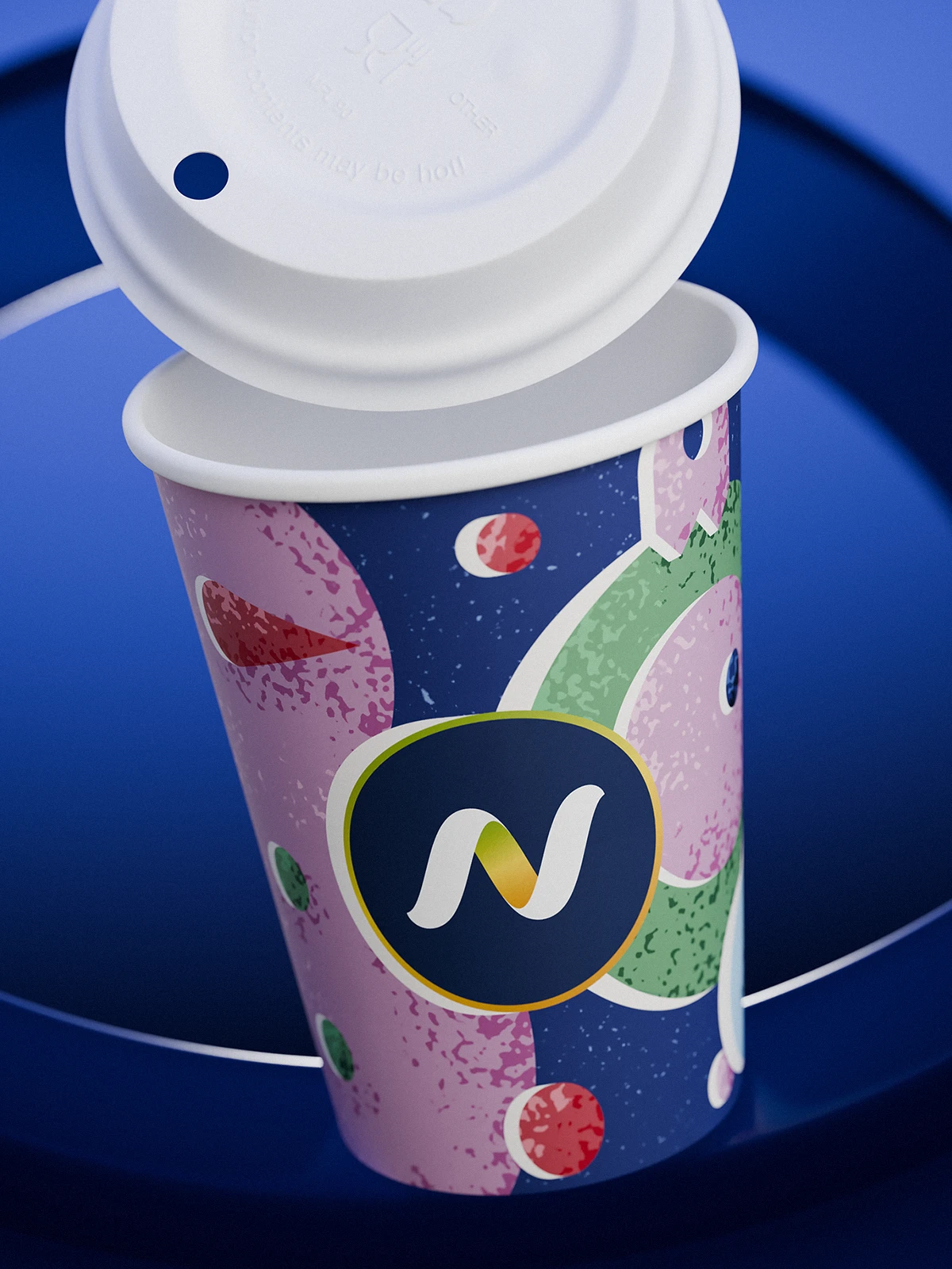 Narvesen Christmas edition paper cup (2024) with a white plastic lid. The festive design combines navy blue, pink, green, and red abstract shapes resembling ornaments and holiday motifs. The front features the Narvesen “N” logo inside a dark blue circle with a golden outline. The cup is set against a vibrant blue background with circular graphic elements.