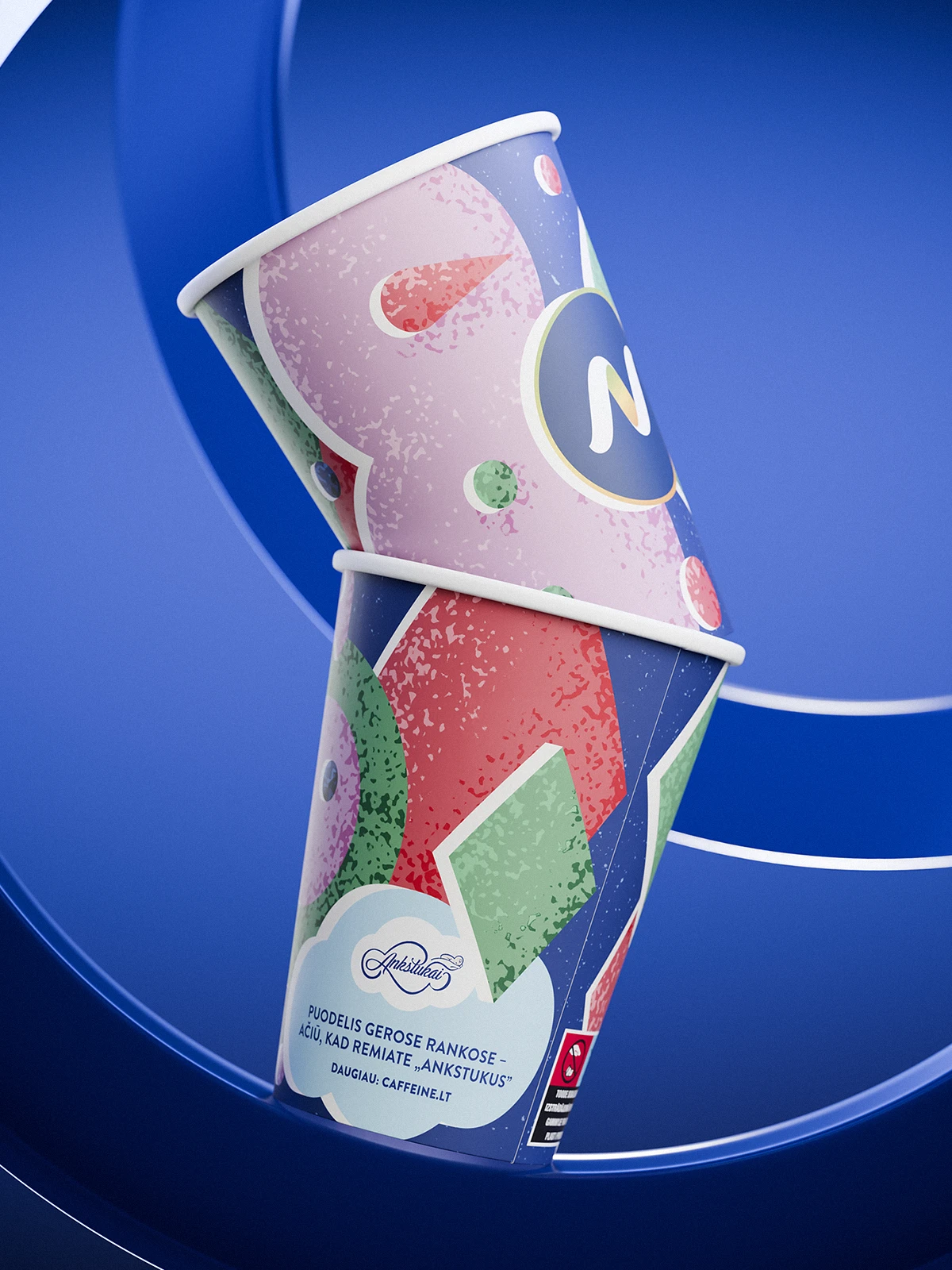 Two stacked Narvesen Christmas edition paper cups (2024) with festive abstract illustrations in pink, red, green, and purple tones. The design features stylized ornaments and shapes, with the Narvesen “N” logo in a dark circle outlined in gold. A white cloud-shaped element at the bottom contains Lithuanian text about supporting the charity “Ankstukai.” The background is a vivid blue with circular graphic elements.