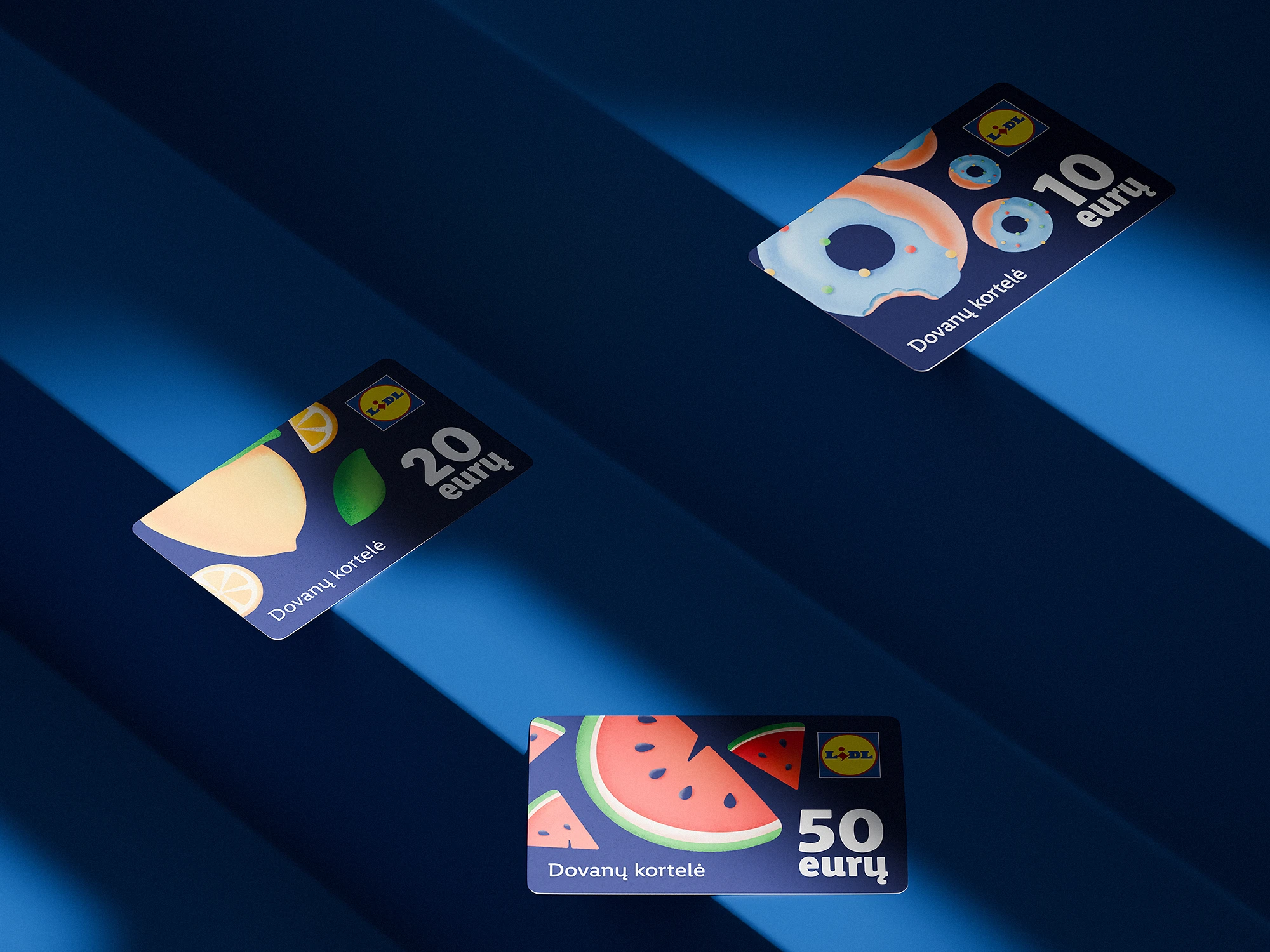 Three Lidl gift cards with playful fruit and donut illustrations lying on a deep blue surface with dramatic shadows. Each card displays a different value in euros—10, 20, and 50—and includes the Lithuanian text “Dovanų kortelė.”