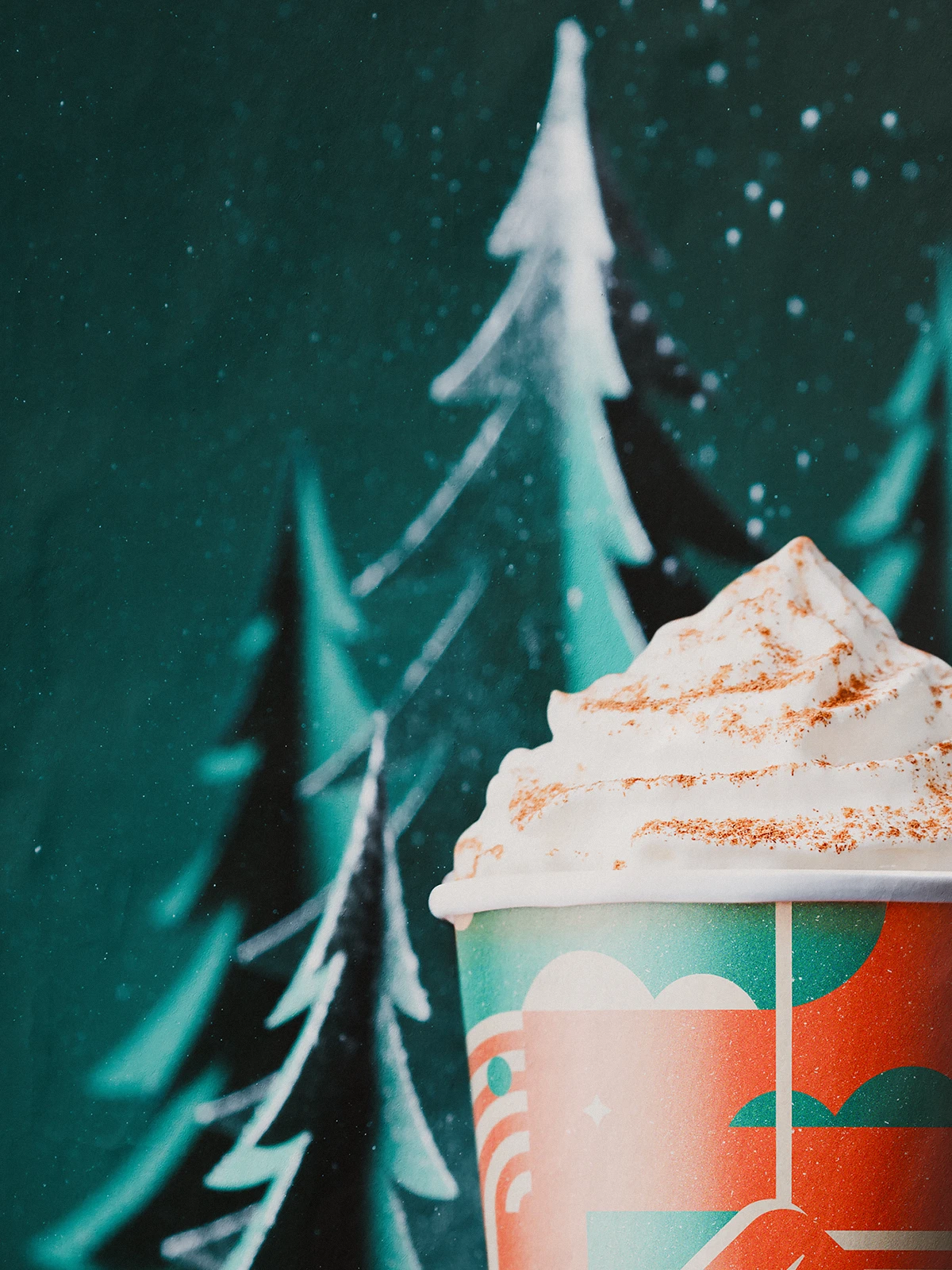 Close-up of Caffeine’s 2021 Christmas cup with whipped cream topping, set in front of paper-cut pine trees and falling snow.