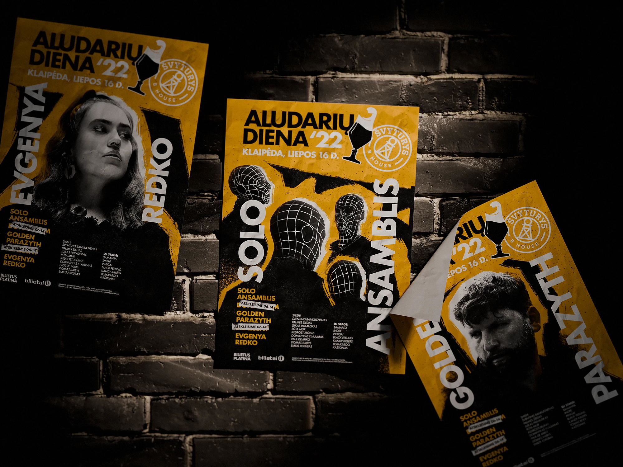 Three music festival posters for Aludarių Diena ’22 displayed on a dark brick wall. The yellow-and-black posters feature portraits of performers Evgenya Redko, Solo Ansamblis, and Golden Parazyth, combining bold typography with graphic wireframe head elements and the Švyturys House logo.