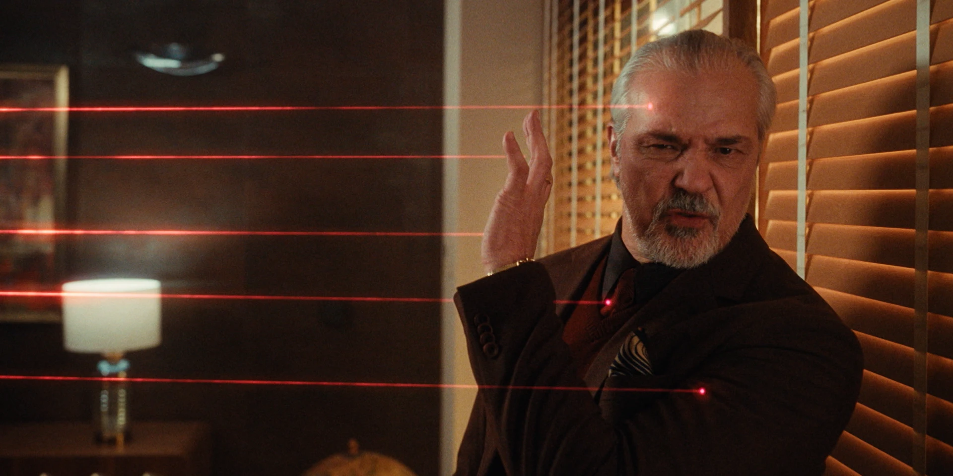 Cinematic shot of an older man in a brown suit inside a dimly lit office, turning sharply as multiple red laser beams cross the room at different heights, warm light from table lamps and window blinds casting dramatic shadows across his face and the wooden interior.