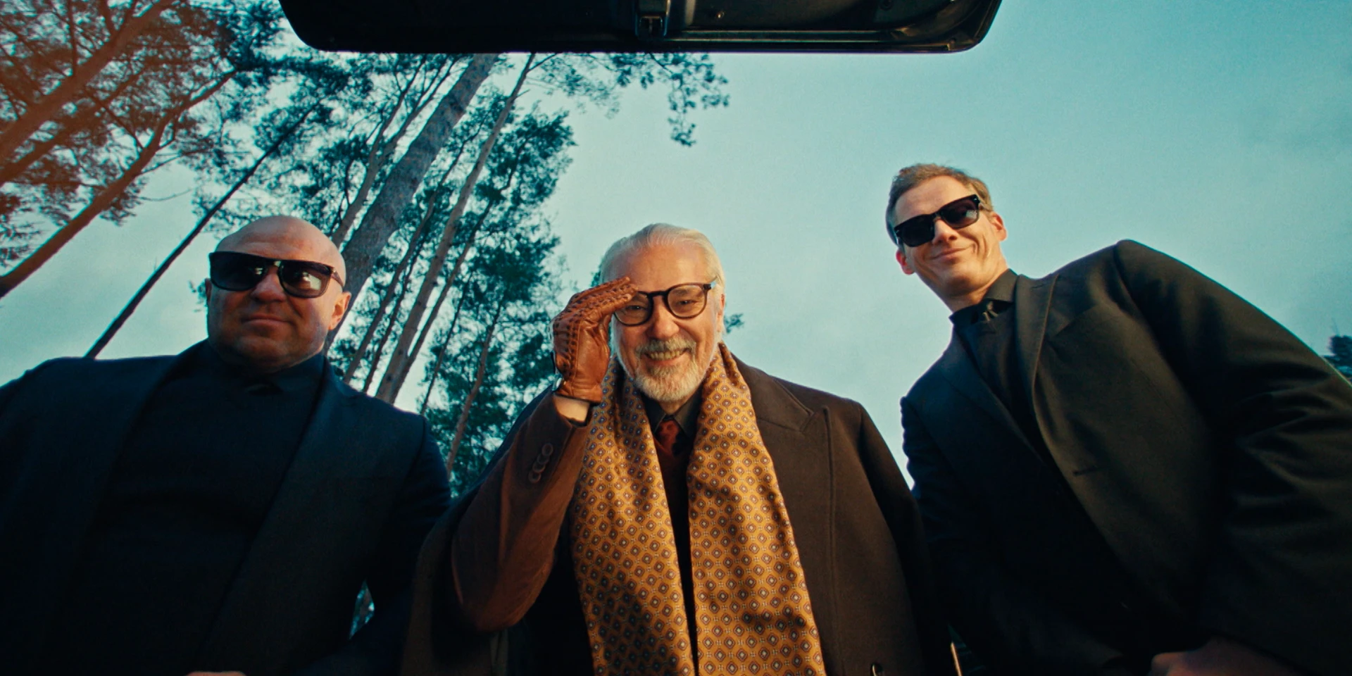 Low-angle cinematic shot from inside an open car trunk, three men in dark suits looking down toward the camera. The central older man smiles confidently, wearing glasses, leather driving gloves and a patterned golden scarf, while the two men beside him wear black sunglasses. Tall pine trees and a pale evening sky rise behind them, creating a dramatic, slightly ironic gangster-style mood.
