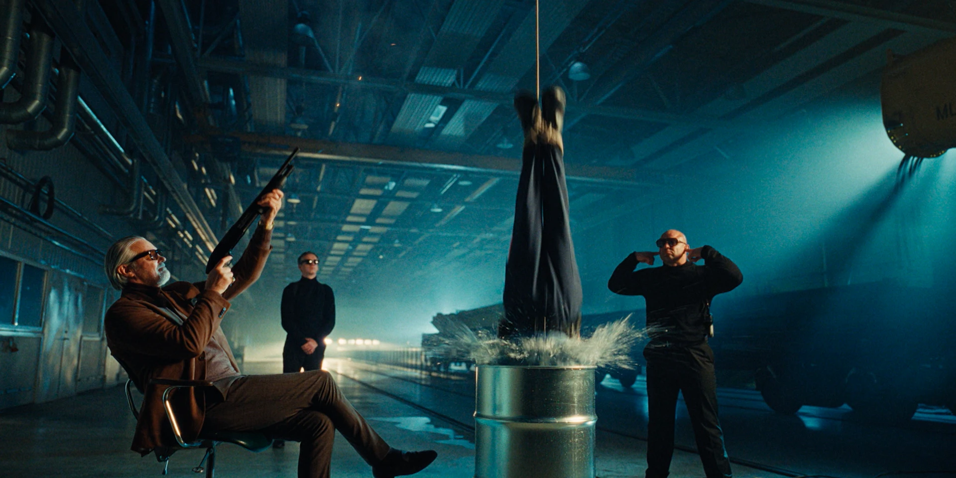 Wide cinematic shot inside a dim industrial warehouse with teal-blue lighting and visible ceiling pipes. On the left, an older man in a brown suit and sunglasses sits casually on a chair, aiming a shotgun upward. In the center, a person hangs upside down above a metal barrel, their head submerged in water as it splashes violently. Two men in black suits and sunglasses stand nearby like bodyguards, with a dark vehicle parked in the background, creating a stylized, crime-film atmosphere.