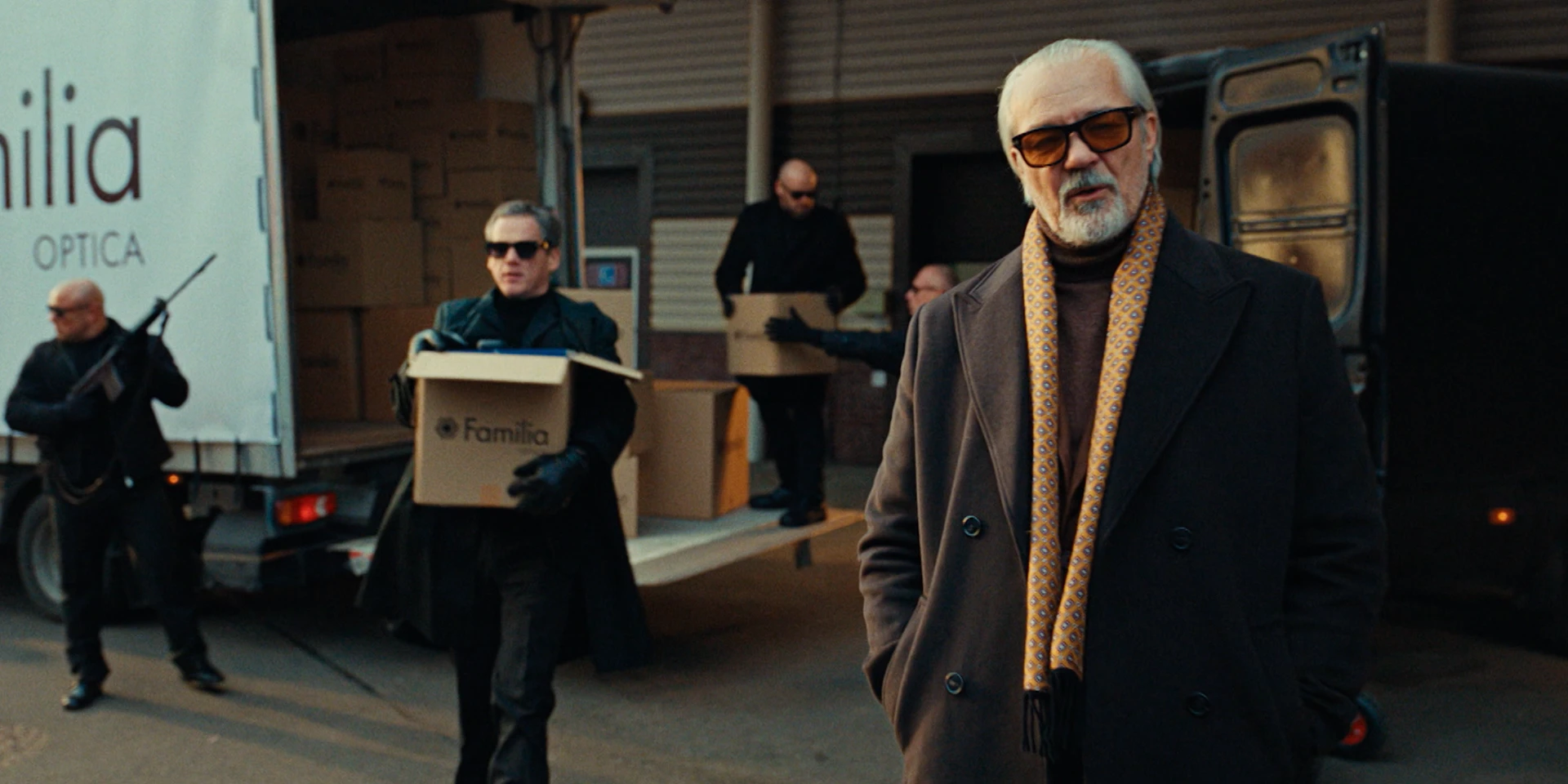 Cinematic outdoor shot in an industrial loading area. In the foreground, an older man with white hair and a beard stands confidently in a dark overcoat and patterned scarf, wearing tinted glasses. Behind him, several men dressed in black with sunglasses unload cardboard boxes labeled “Familia” from the back of a truck. One man stands guard holding a firearm, reinforcing the stylized crime-film aesthetic of the scene.