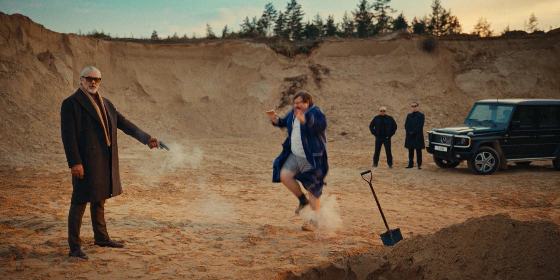 Wide cinematic shot in a sandy quarry at dusk. On the left, an older man in a long dark coat and sunglasses fires a handgun toward the ground, smoke rising from the barrel. In the center, a frightened man in a bathrobe and slippers jumps in panic near a freshly dug pit, kicking up sand. A shovel stands planted beside the hole. In the background, two men in black suits and sunglasses stand watching next to a black SUV, reinforcing the stylized, darkly comic crime-film atmosphere.