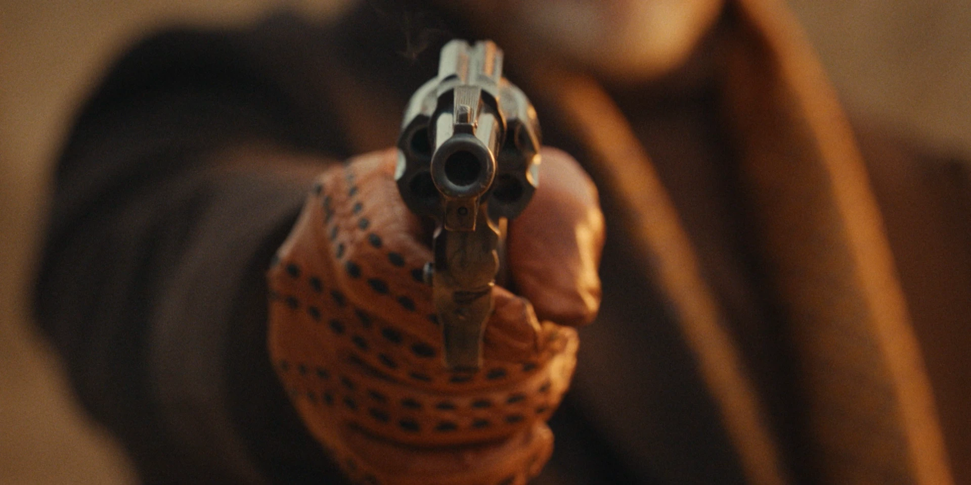 Extreme close-up shot of a revolver aimed directly at the camera. The barrel dominates the frame, centered and in sharp focus, while the man holding it remains blurred in the background. He wears a brown leather driving glove, and the warm, golden light enhances the tense, cinematic atmosphere.