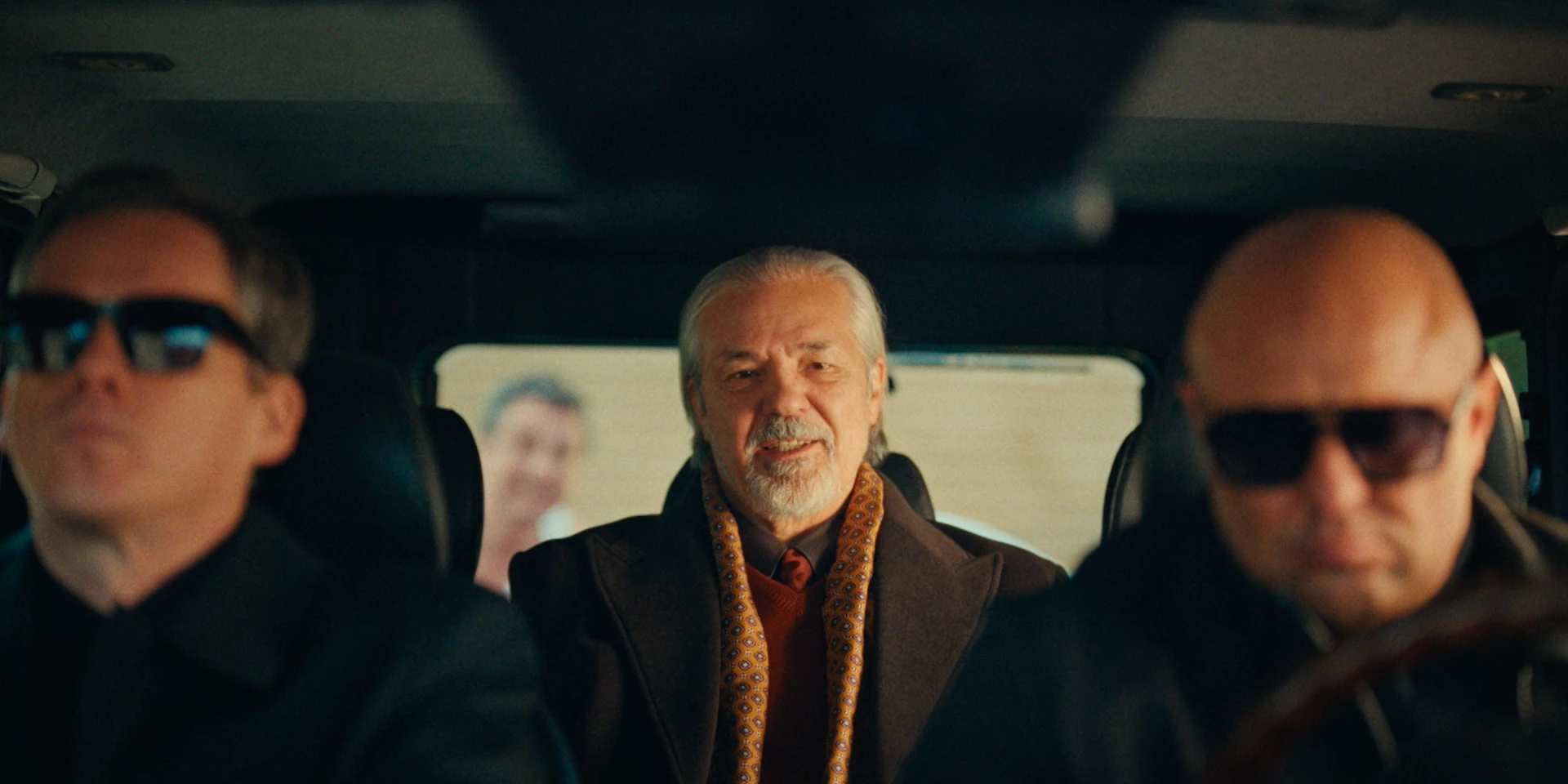 Medium shot from inside a car, camera positioned on the dashboard facing the back seat. In the foreground, slightly out of focus, sit two men in black suits and dark sunglasses — one driving, one in the passenger seat. In sharp focus in the center of the back seat is an older man with silver hair and a trimmed beard. He wears a dark coat with a patterned golden-brown scarf and looks forward with a calm, confident expression. Through the rear window behind him, a blurred figure stands outside, adding subtle tension. The lighting is warm and cinematic, with soft contrast and a composed, gangster-film atmosphere.