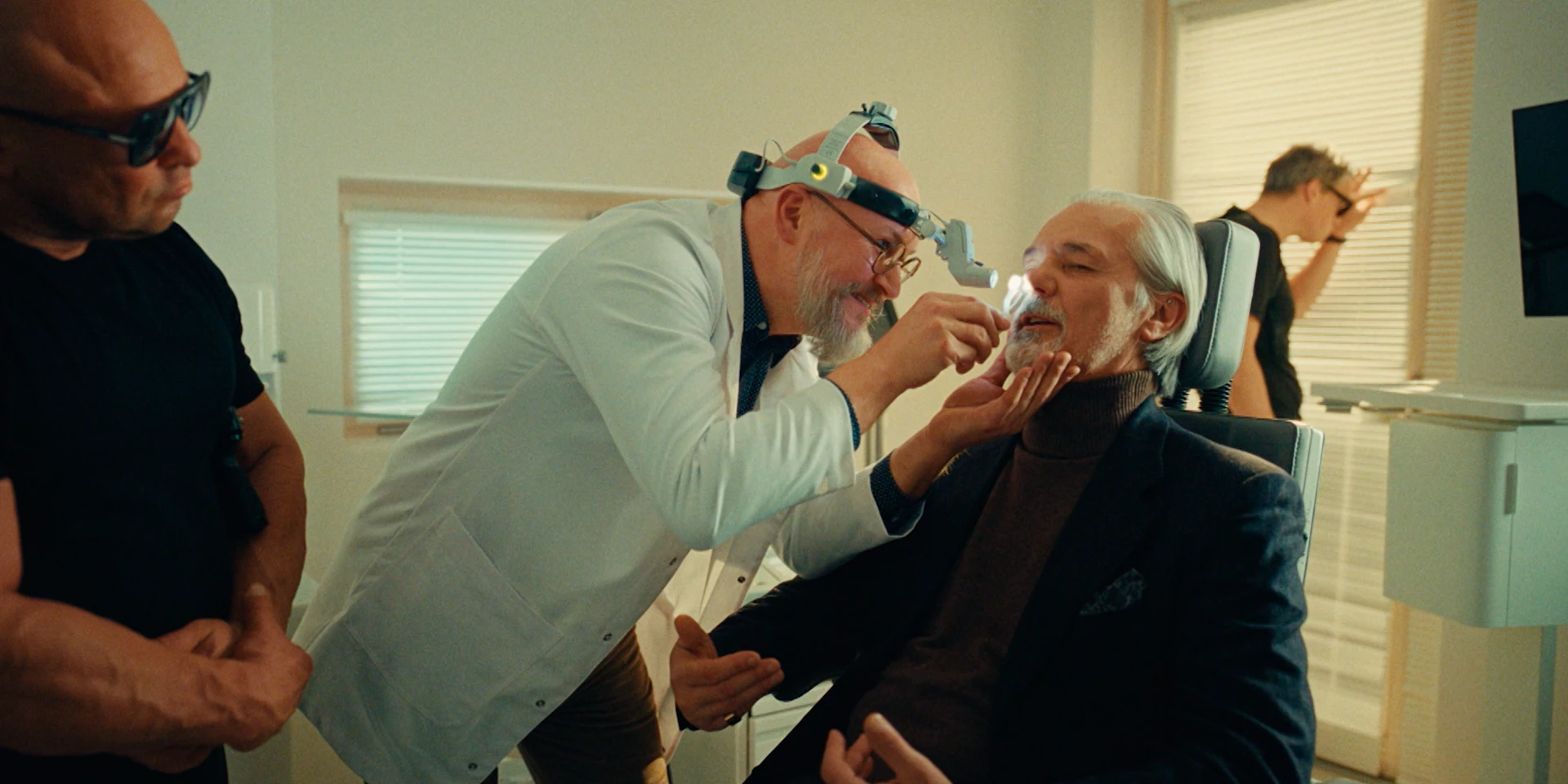 Wide interior shot of a bright clinic. In the center, an older silver-haired man in a dark blazer and turtleneck sits in a dental chair, slightly reclined, composed but mildly uncomfortable. A dentist in a white coat and headlamp leans in close, carefully examining his teeth with dental tools, one hand gently holding the patient’s chin. On the left, a muscular bodyguard in a black T-shirt and sunglasses stands with arms folded, watching seriously. In the background near the window blinds, another suited man stands turned away, hand to his forehead, as if stressed or impatient. The lighting is warm and cinematic despite the clinical setting, creating a contrast between the sterile medical environment and the tense, gangster-like entourage atmosphere.
