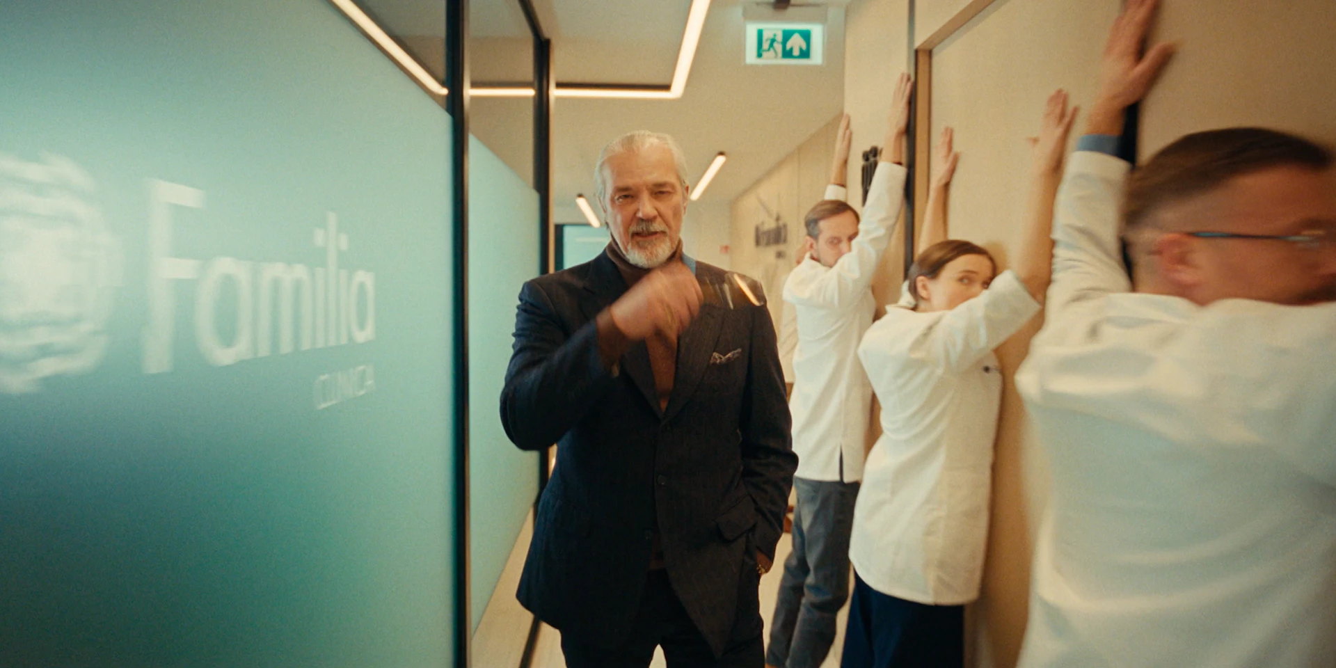 Wide hallway shot inside a modern clinic. On the left, a frosted glass wall with the Familia Clinica logo runs along the corridor. The lighting is clean and warm, with minimalistic ceiling strips guiding perspective toward the back. In the center foreground, a silver-haired older man in a dark tailored suit walks confidently toward camera, adjusting his glasses with one hand while the other rests casually in his pocket. His expression is calm, controlled, slightly dominant. On the right side of the frame, three medical staff members in white coats stand facing the wall with their hands raised above their heads, as if being searched or detained. One of them glances sideways with visible anxiety. The tone remains cinematic and slightly ironic — a polished medical environment contrasted with exaggerated “crime-scene” body language, maintaining the gangster parody atmosphere.