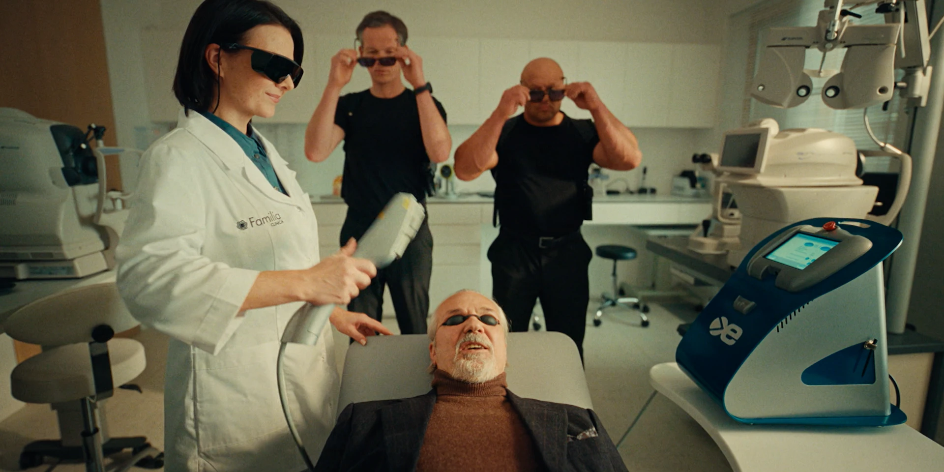 Clinical treatment room, bright and sterile. Foreground: an older silver-haired man in a dark suit lies back in a medical chair, wearing small protective eye covers. His expression is tense but controlled — trying to maintain dignity. Left: a female doctor in a white Familia Clinica coat holds a medical laser device, angled toward his face. She wears dark protective glasses and looks confident and composed, slightly amused. Background: two large bodyguard-type men in black T-shirts simultaneously put on protective glasses, as if preparing for something dramatic. Their synchronized movement adds subtle humor. Right side: modern aesthetic medical equipment with a digital screen and blue accents reinforces the high-tech environment. The overall mood stays cinematic and ironic — gangster energy transported into a polished aesthetic clinic setting, exaggerating the seriousness of a simple cosmetic procedure.